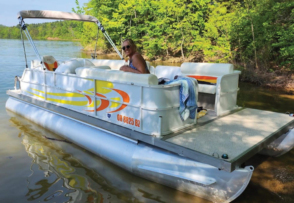 A smiling woman relaxing on a white and orange pontoon boat floating along a tree-lined shoreline.