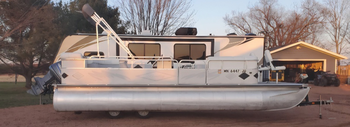 Side profile of a grey pontoon boat parked in front of a large camper trailer and garage.