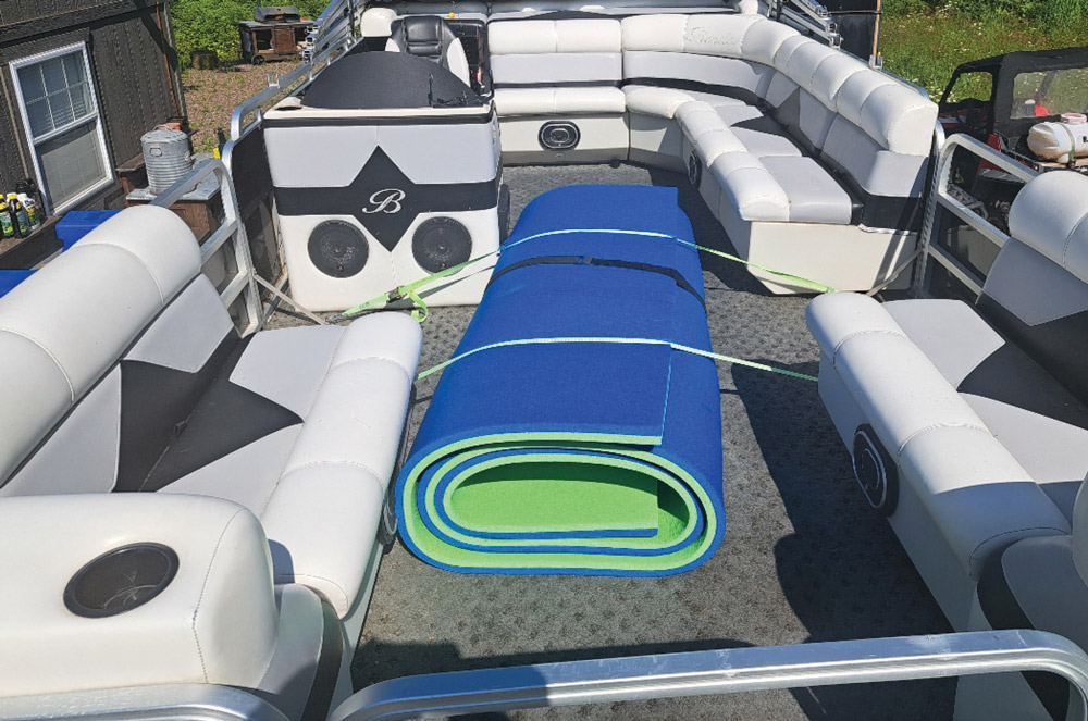 Interior deck of a pontoon boat featuring grey vinyl seating and a large rolled-up blue floating water mat.