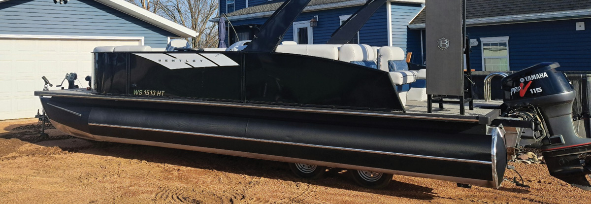 Side view of a restored black Bentley pontoon boat parked on dirt next to a blue garage.