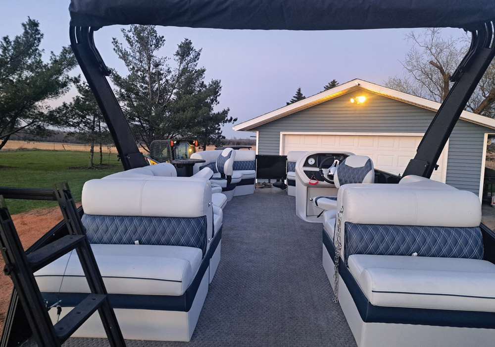 Clean interior of a pontoon boat featuring light grey seats with blue diamond stitching, parked near a garage at dusk.
