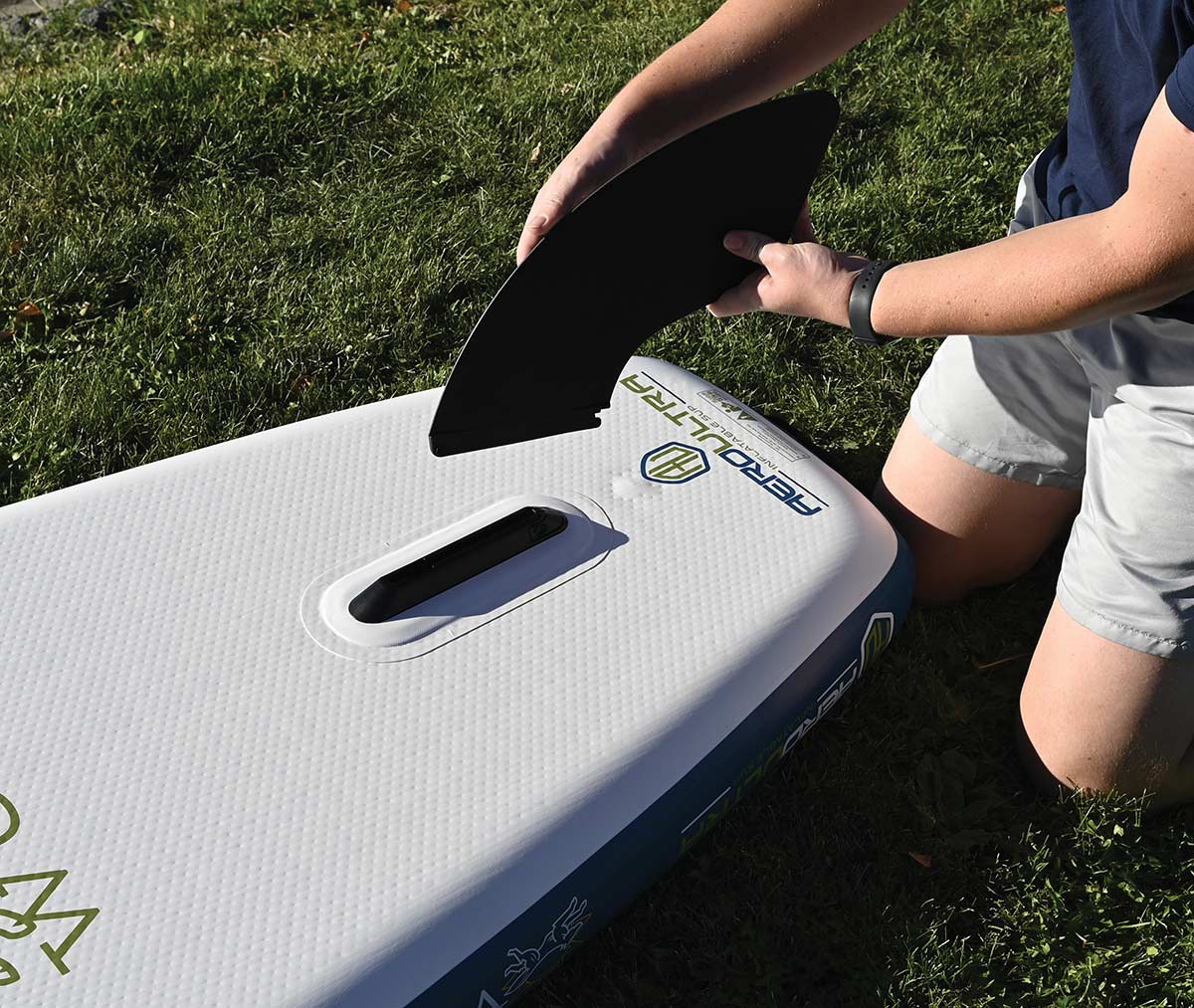 Person attaching a black removable fin into the slot of an inflatable paddle board.