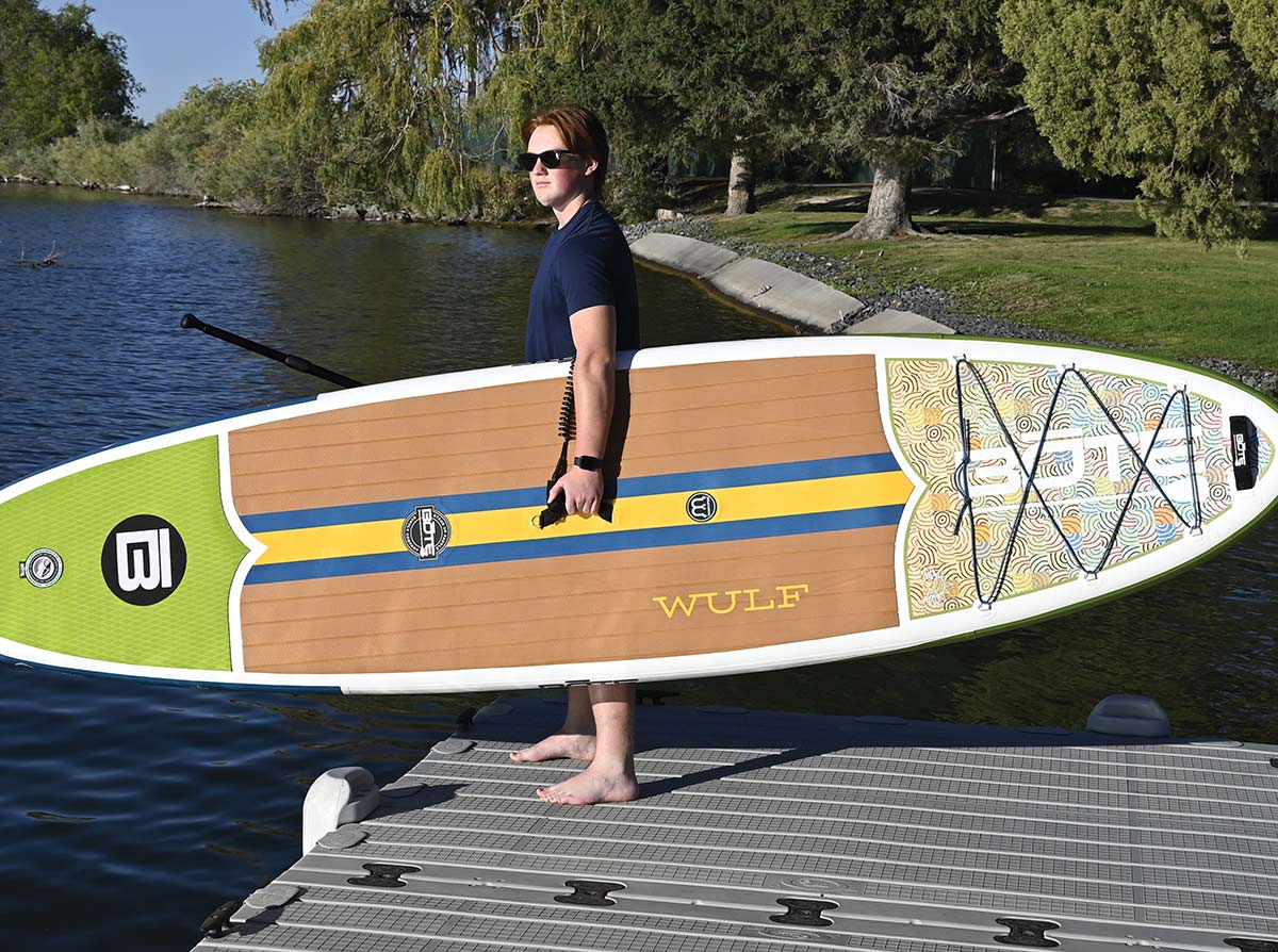 Person holding a BOTE Wulf inflatable paddle board vertically while standing on a dock.