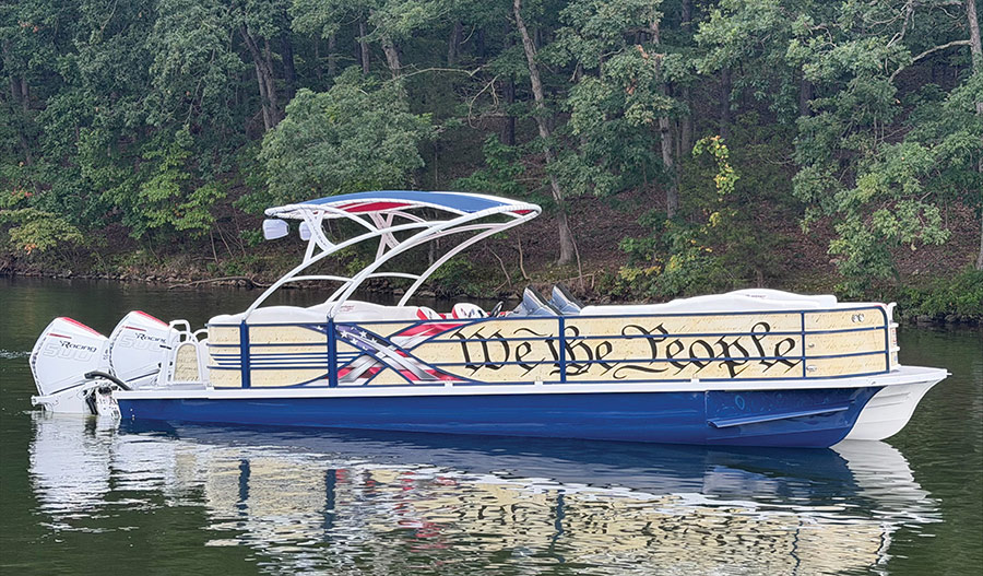 A pontoon boat decorated with "We the People" script and American flag graphics anchored near a forested shoreline.