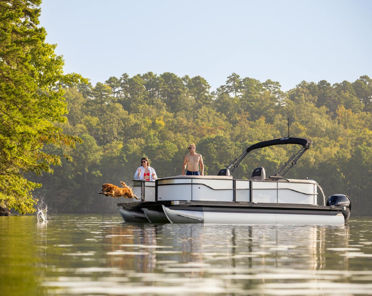 Golden retriever leaping off the front deck of a pontoon boat into a lake while two people watch.