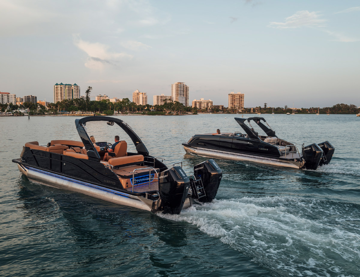 Two luxury Harris pontoon boats cruising near a coastal city skyline at dusk.