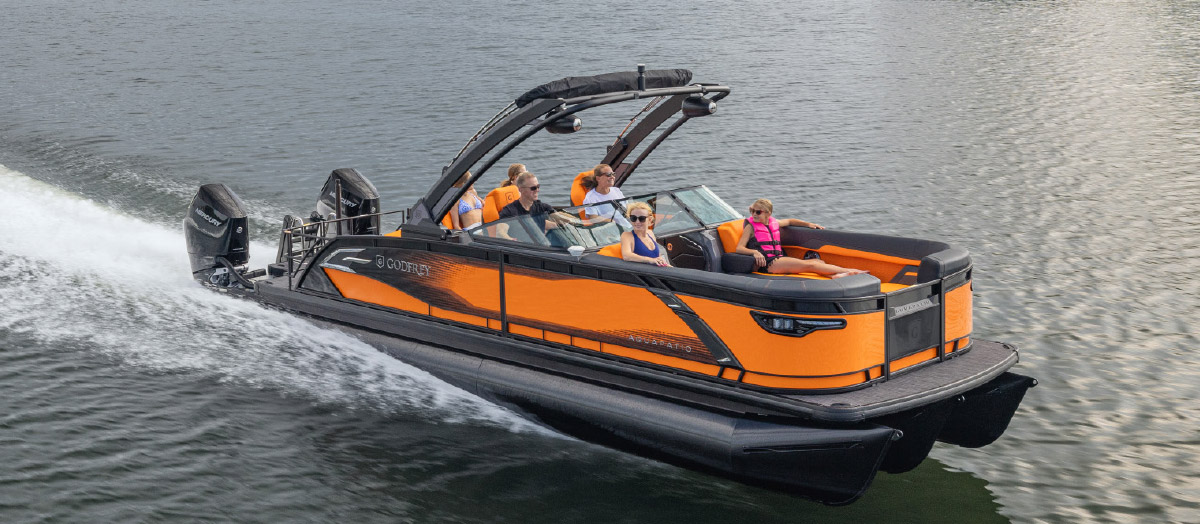 Bright orange and black Godfrey pontoon boat speeding across open water with passengers.