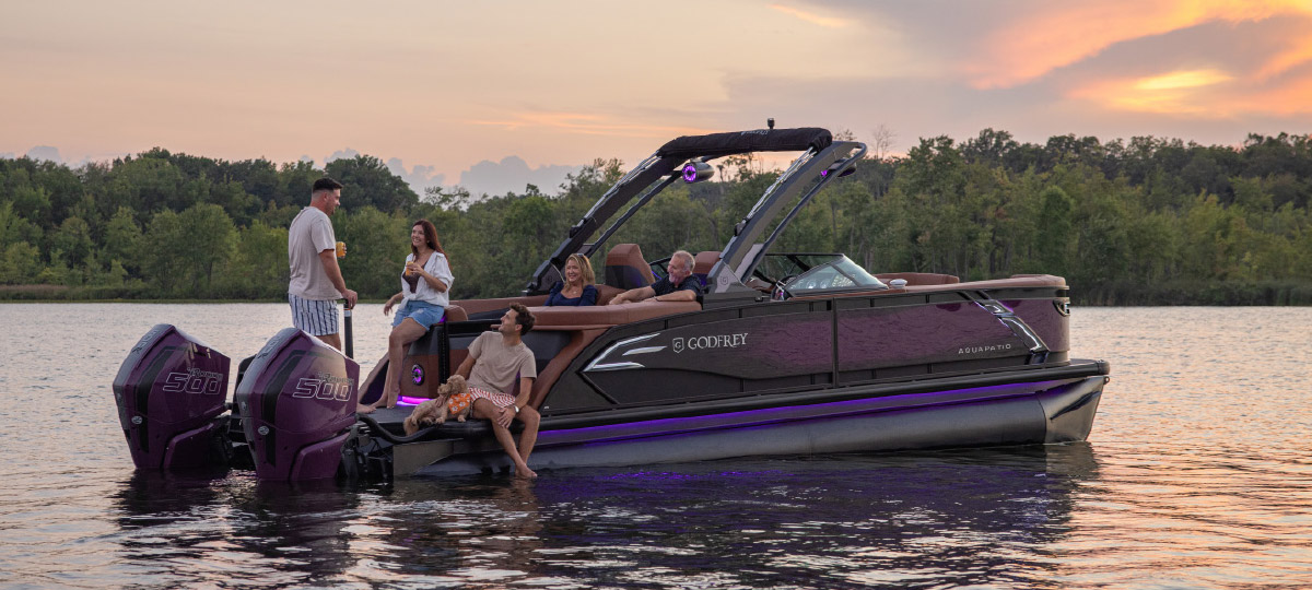Group of friends relaxing on a purple-lit Godfrey pontoon boat equipped with twin 500 hp engines at sunset.