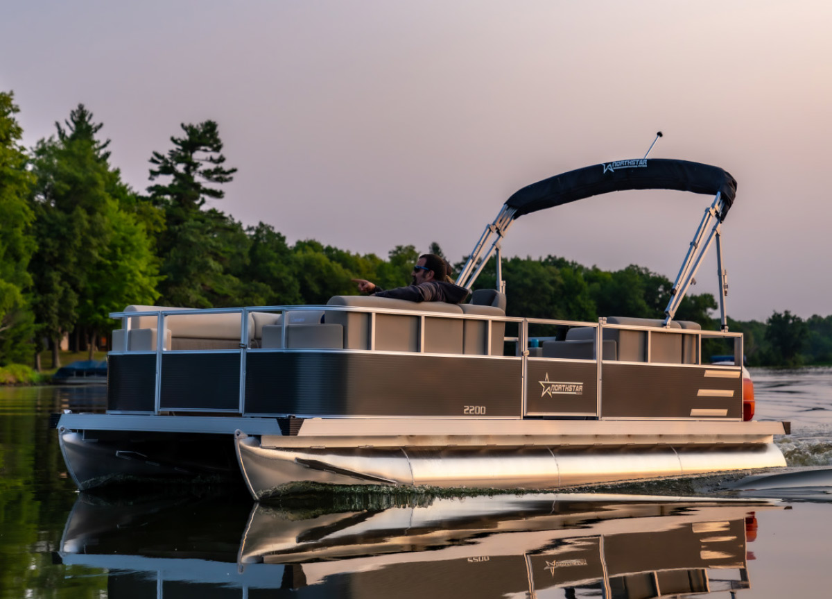 Northstar 2200 pontoon boat anchored on calm water near a forested shoreline at dusk.