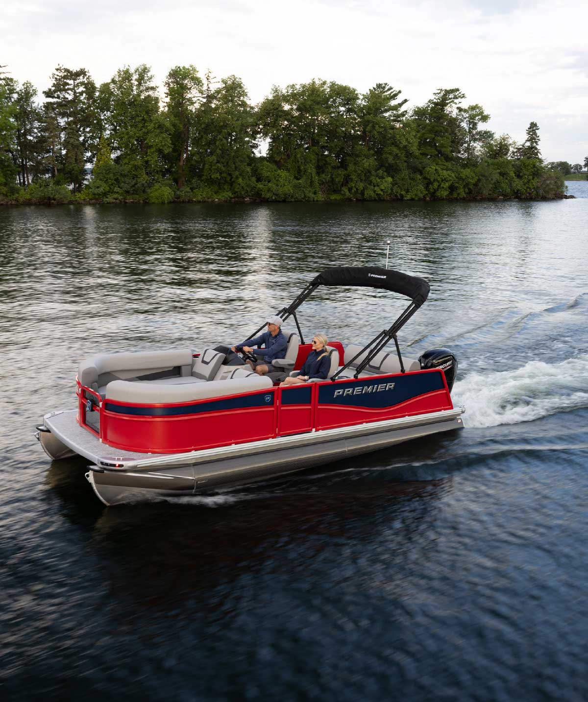 Red and navy Premier pontoon boat cruising past a tree-lined shore with two people on board.