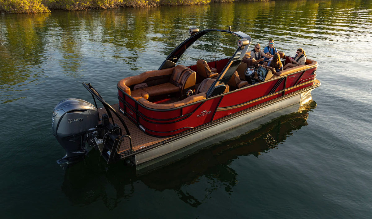 High-angle view of a red and tan SunTracker pontoon boat with passengers idling near a wooded bank.