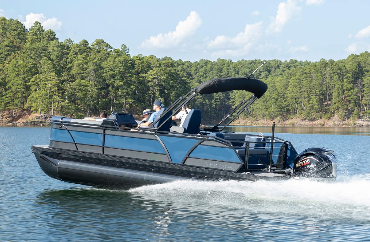 Side view of a blue and black Veranda pontoon boat cruising along a lake near a pine-covered shoreline.