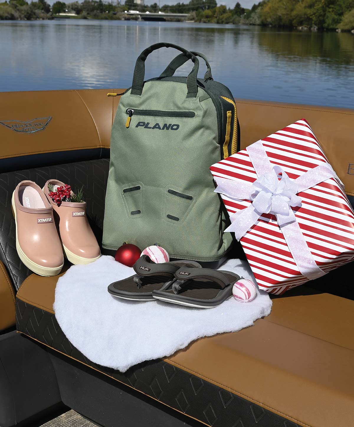 A green Plano backpack, pink Xtratuf clogs, dark Huk flip-flops, and a wrapped present displayed with holiday ornaments on a boat seat.