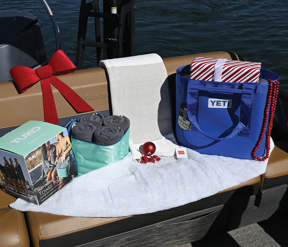 A blue Yeti tote bag holding a wrapped gift, a Tuko towel pack, and a white blanket displayed with holiday decorations on a pontoon boat seat.