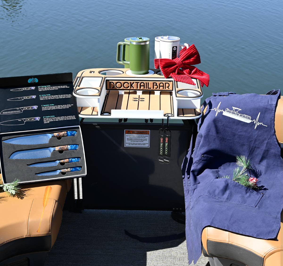 A Docktail Bar organizer holding insulated mugs, a boxed set of blue chef knives, and a navy apron with a pontoon graphic displayed on a boat seat with holiday decor.
