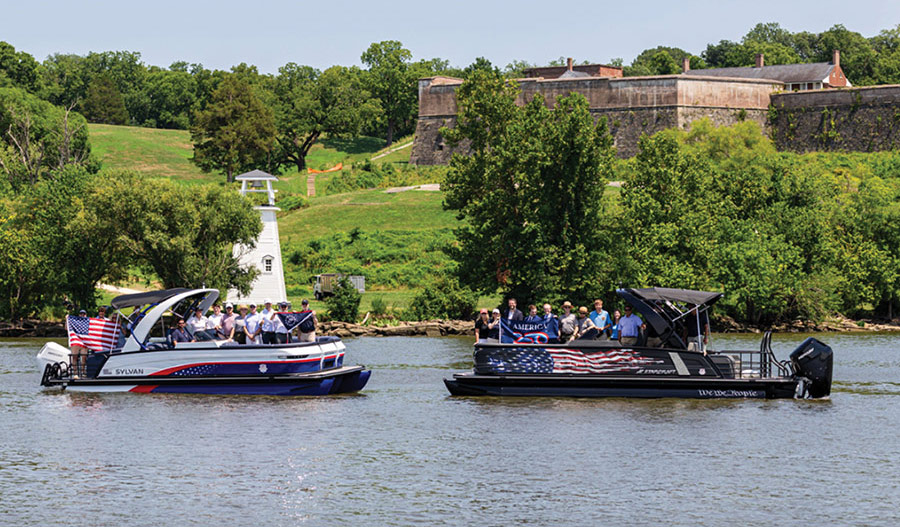 Two patriotic-themed pontoon boats carrying passengers past a historic brick fort and lighthouse.